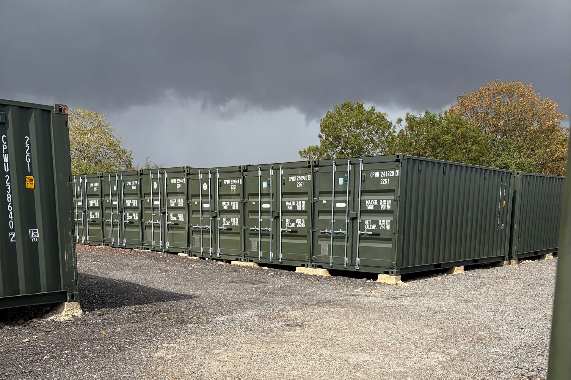 Row of green storage containers under a stormy sky, with trees behind